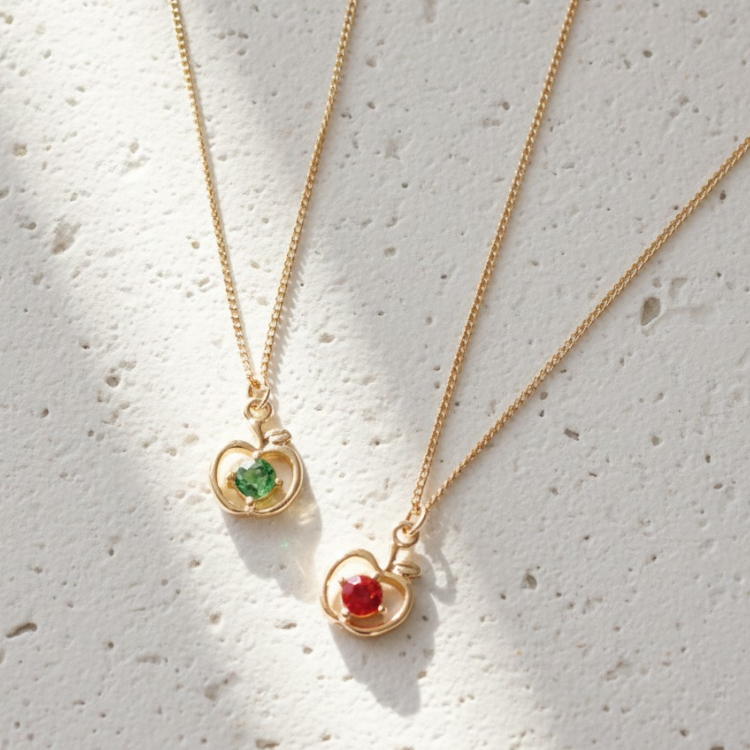 Two gold necklaces with heart-shaped pendants featuring green and red gemstones on a light gray background.