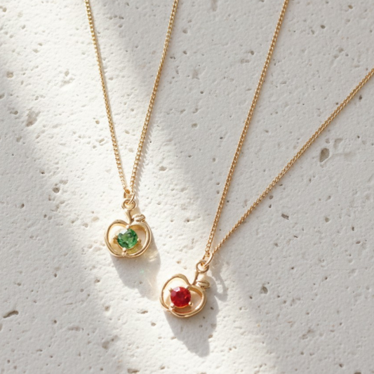 Two gold necklaces with heart-shaped pendants featuring green and red gemstones on a light gray background.