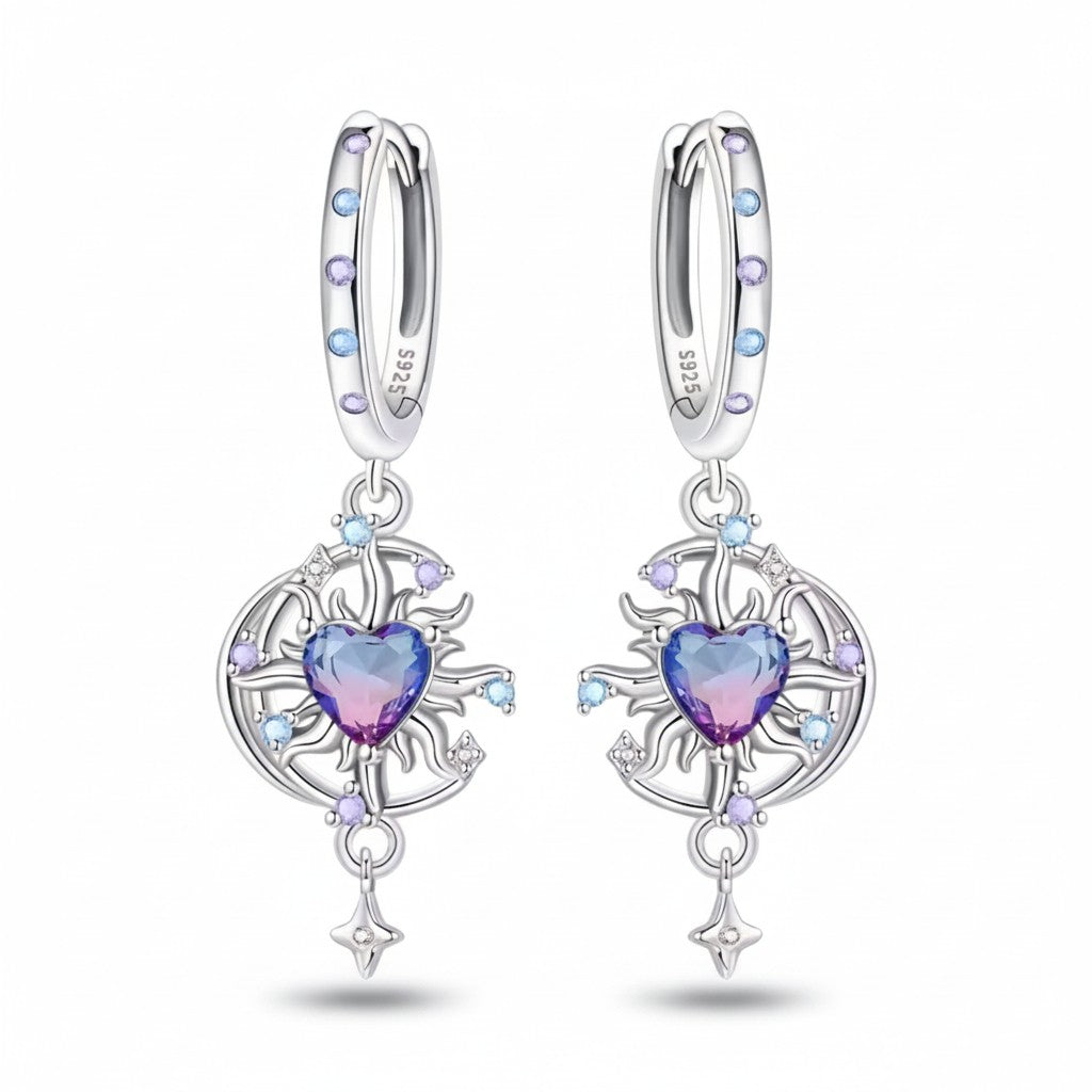 A pair of sterling silver huggie hoop earrings with dangling circular filigree charms. Each earring features a central heart-shaped crystal that transitions from purple to pink, surrounded by small light blue and lavender crystals, on a clean white background.