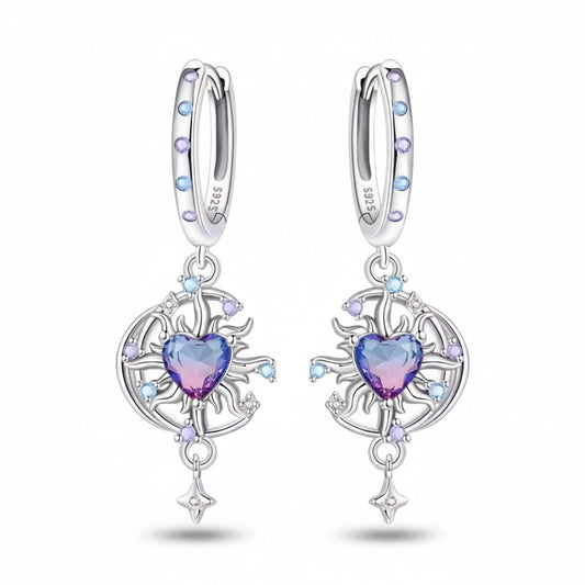 A pair of sterling silver huggie hoop earrings with dangling circular filigree charms. Each earring features a central heart-shaped crystal that transitions from purple to pink, surrounded by small light blue and lavender crystals, on a clean white background.