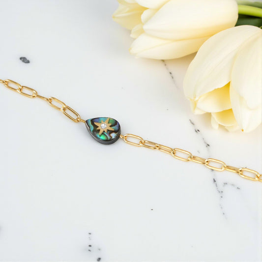 Gold bracelet with a heart-shaped abalone shell charm on a marble surface with white tulips.