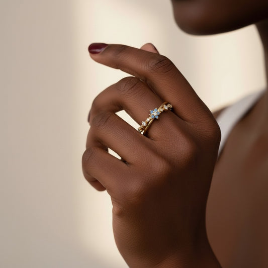 A close-up of a person's hand wearing a delicate gold ring with small clear stones and a central light blue flower-shaped accent, set against a soft beige background.