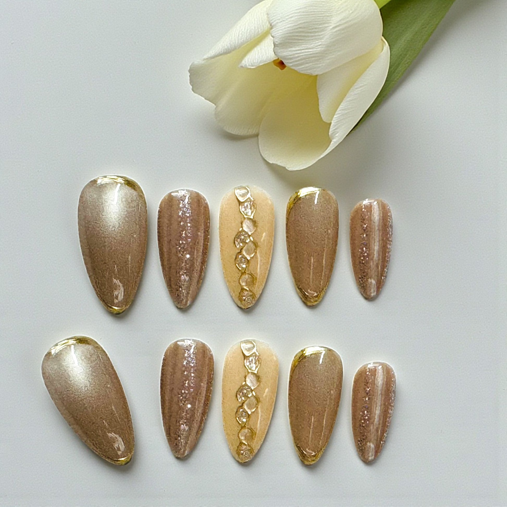 Set of gold and bronze nail art designs on a white surface with a white tulip.