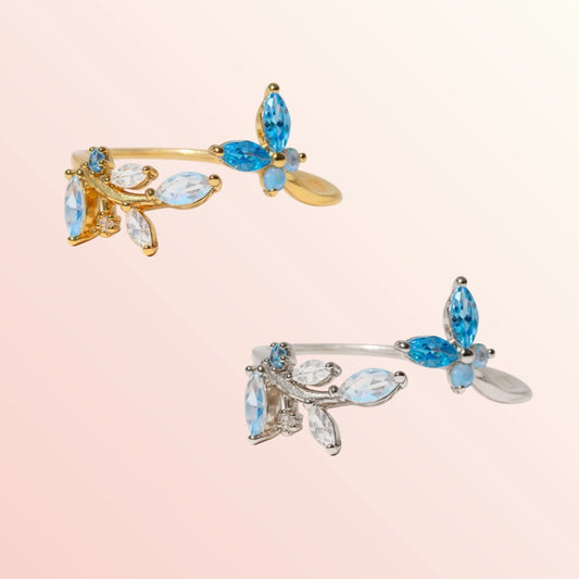 Two butterfly-shaped jewelry pieces with blue and clear gemstones on a light pink background