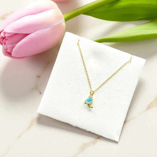 Gold necklace with a blue pendant on a white card, next to a pink tulip on a marble surface.