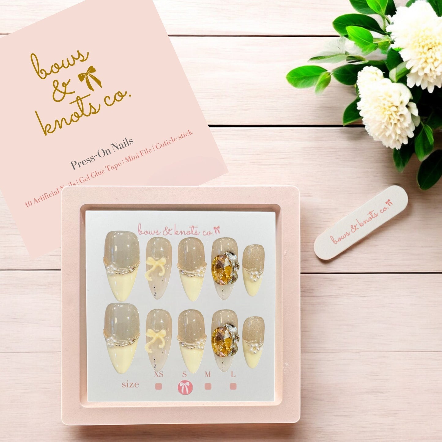 Press-on nails set with decorative designs on a pink card, accompanied by a small white flower and green leaves on a wooden surface.