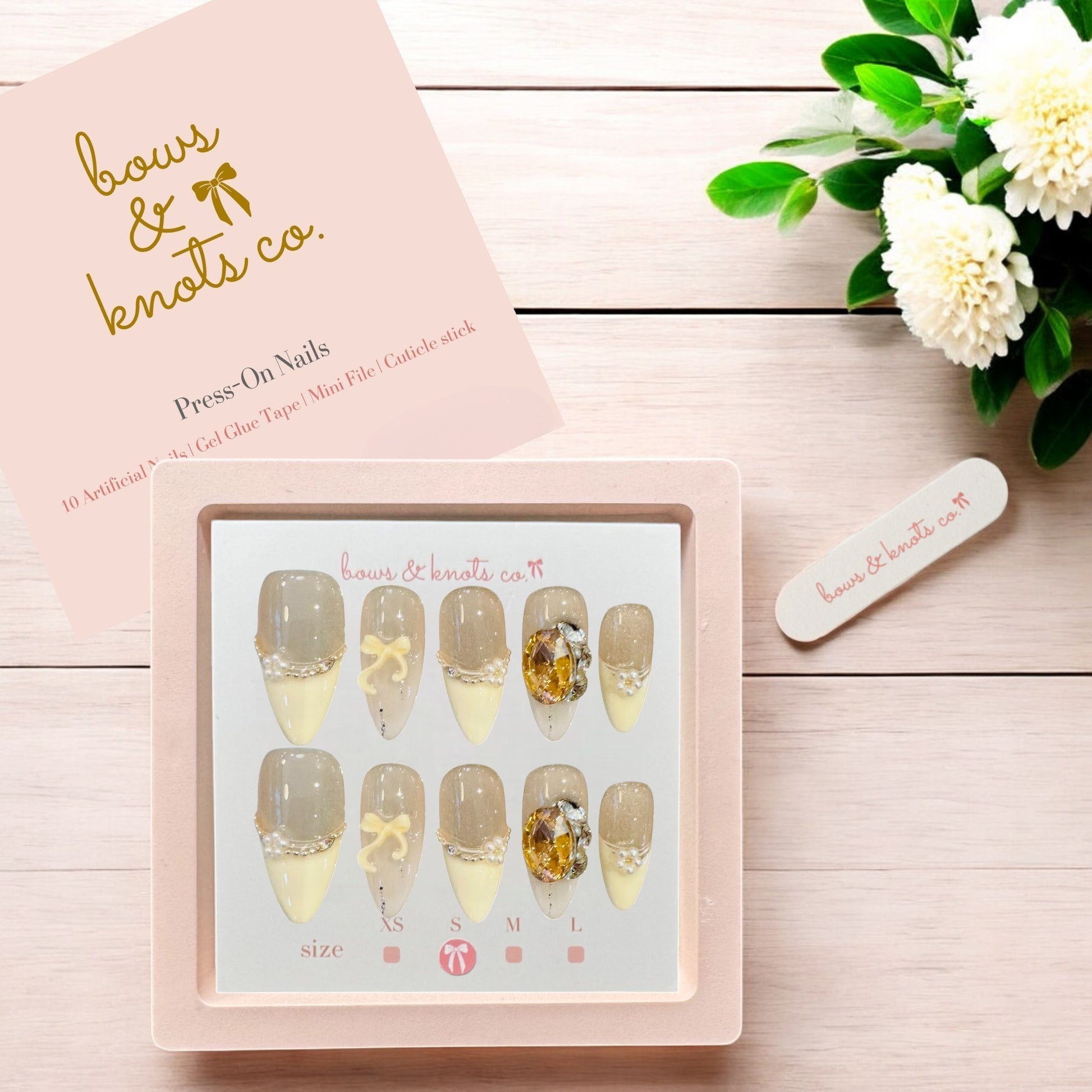 Press-on nails set with decorative designs on a pink card, accompanied by a small white flower and green leaves on a wooden surface.