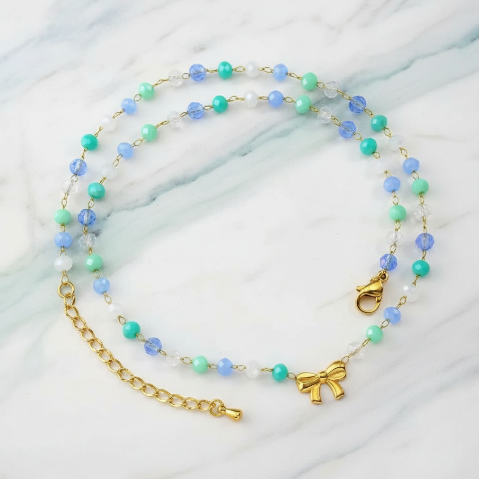 Necklace with blue and green beads and a gold bow charm on a marble background