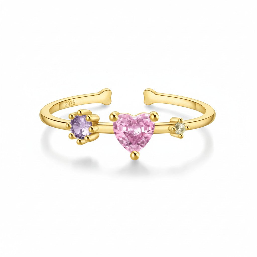 A gold-plated open ring band featuring a central heart-shaped pink gemstone, flanked by a smaller purple heart gemstone on the left and a tiny clear heart gemstone on the right. The inner band is marked '925'.
