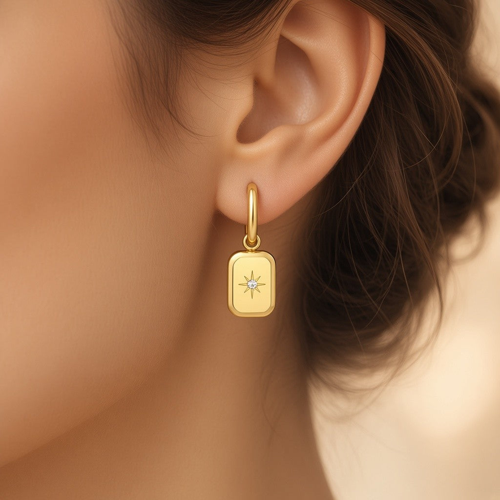 A close-up of a woman's ear wearing a Gold stainless steel hoop earrings with rectangular North Star charms and sparkling center stones.