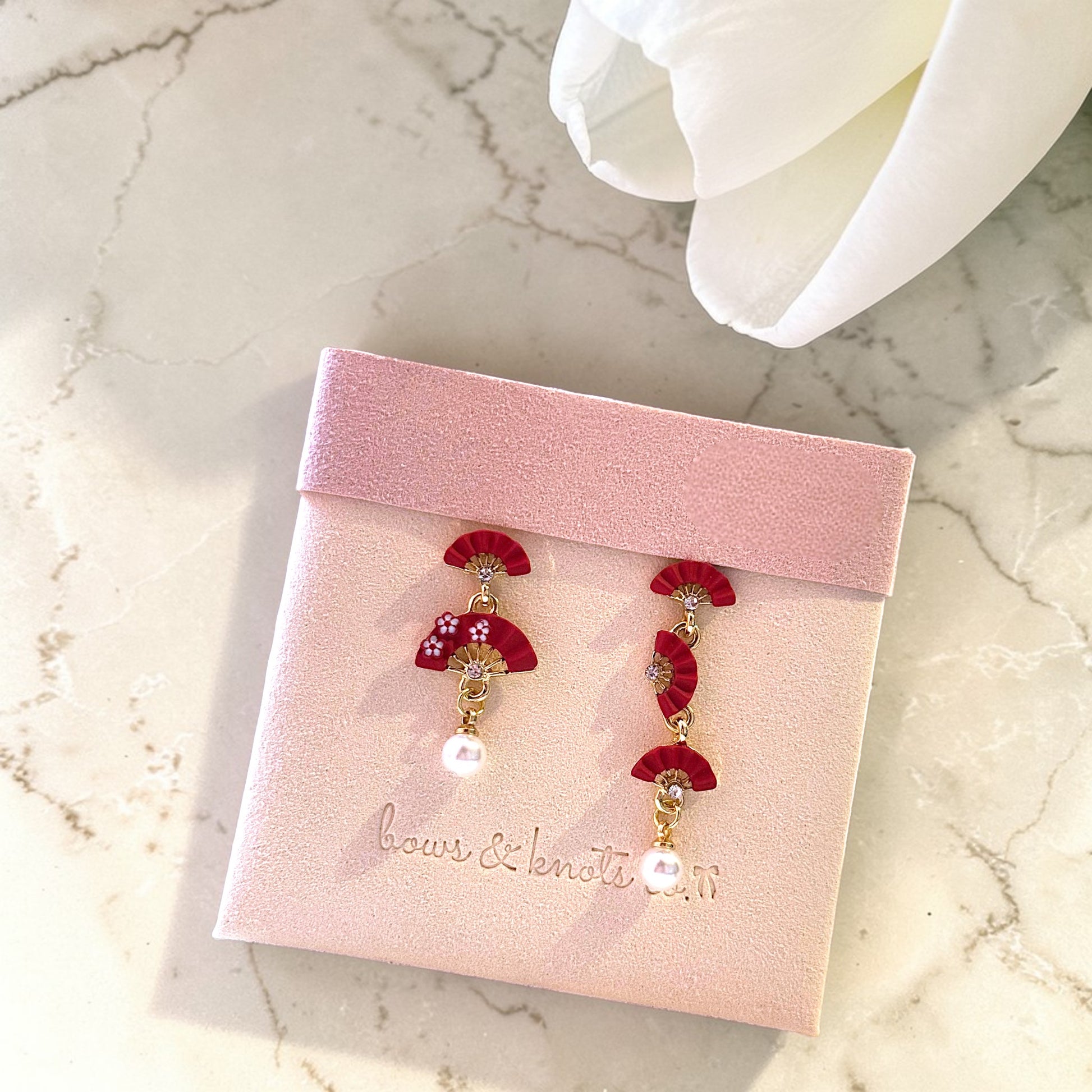 Red floral earrings on a pink jewelry card with 'bows & Sequins' branding on a marble surface.