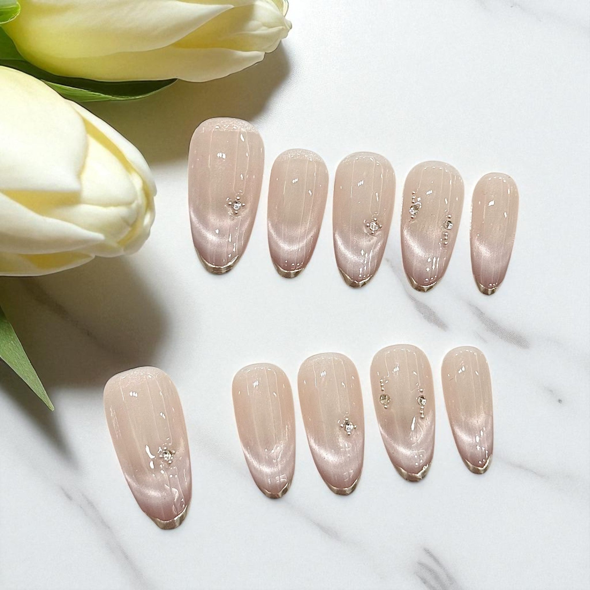 Set of nude-colored nail polish samples with decorative crystals on a white surface with tulips.
