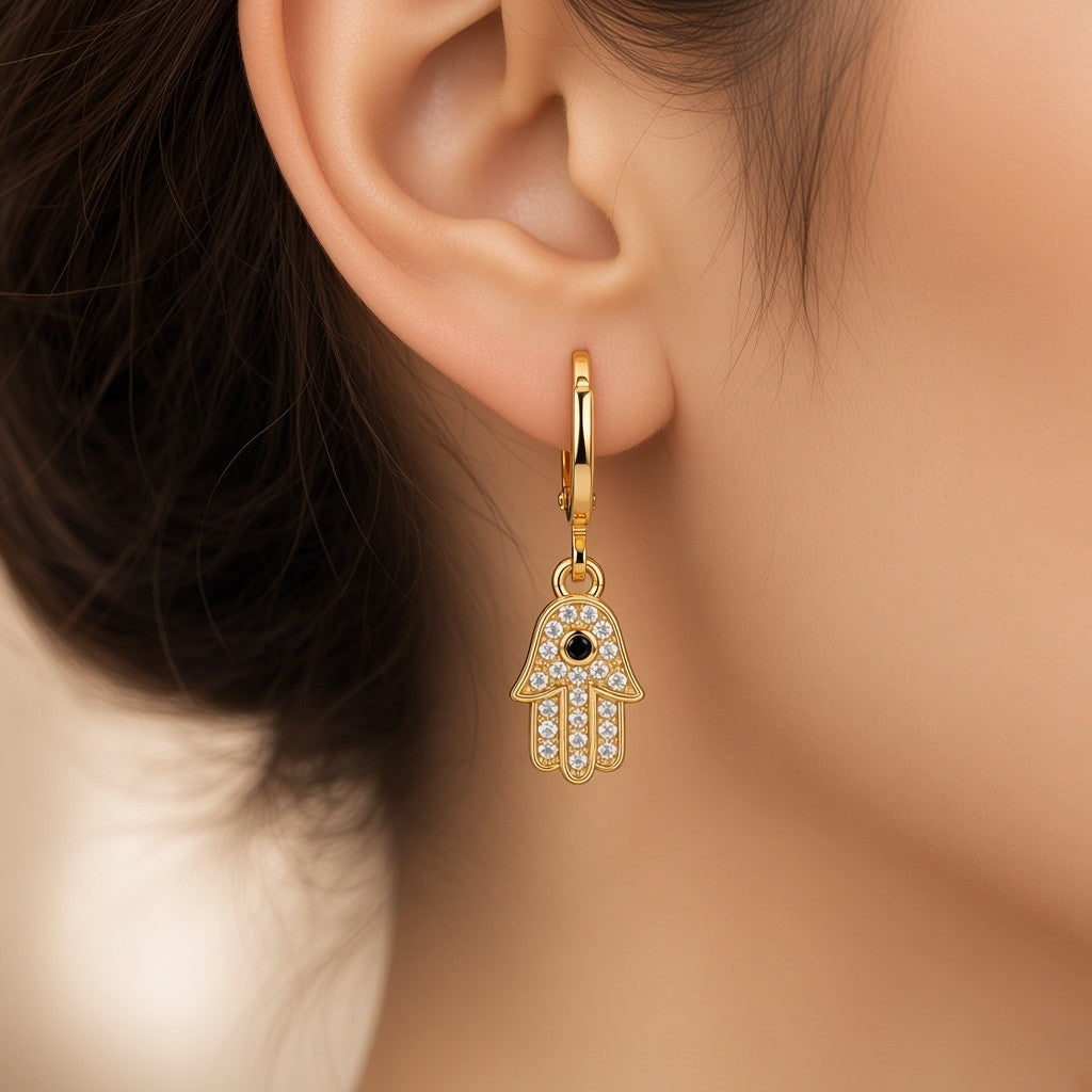A close-up of a model's ear wearing a Gold stainless steel hoop earrings with crystal Hamsa hand charms and black center stones.