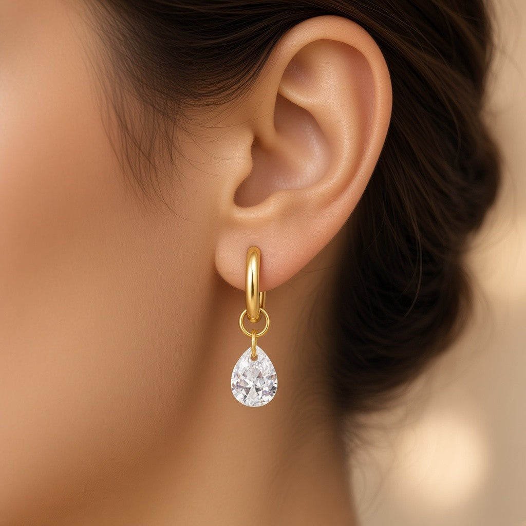A close-up of a model's ear wearing a Gold stainless steel hoop earrings with clear faceted teardrop crystal charms.