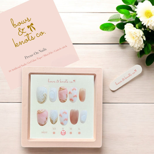 Set of press-on nails with a 'bows & knots co.' box on a wooden surface with flowers.
