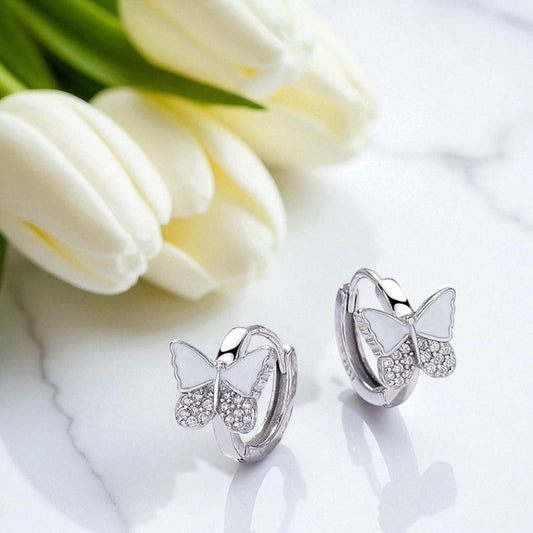 Butterfly-shaped silver earrings with white stones on a white surface with tulips in the background.