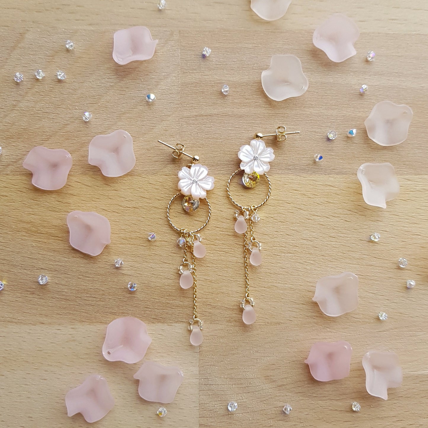 A pair of gold and white floral dangle earrings with pink rose quartz beads, displayed on a light wood surface scattered with pink petals and small clear rhinestones.