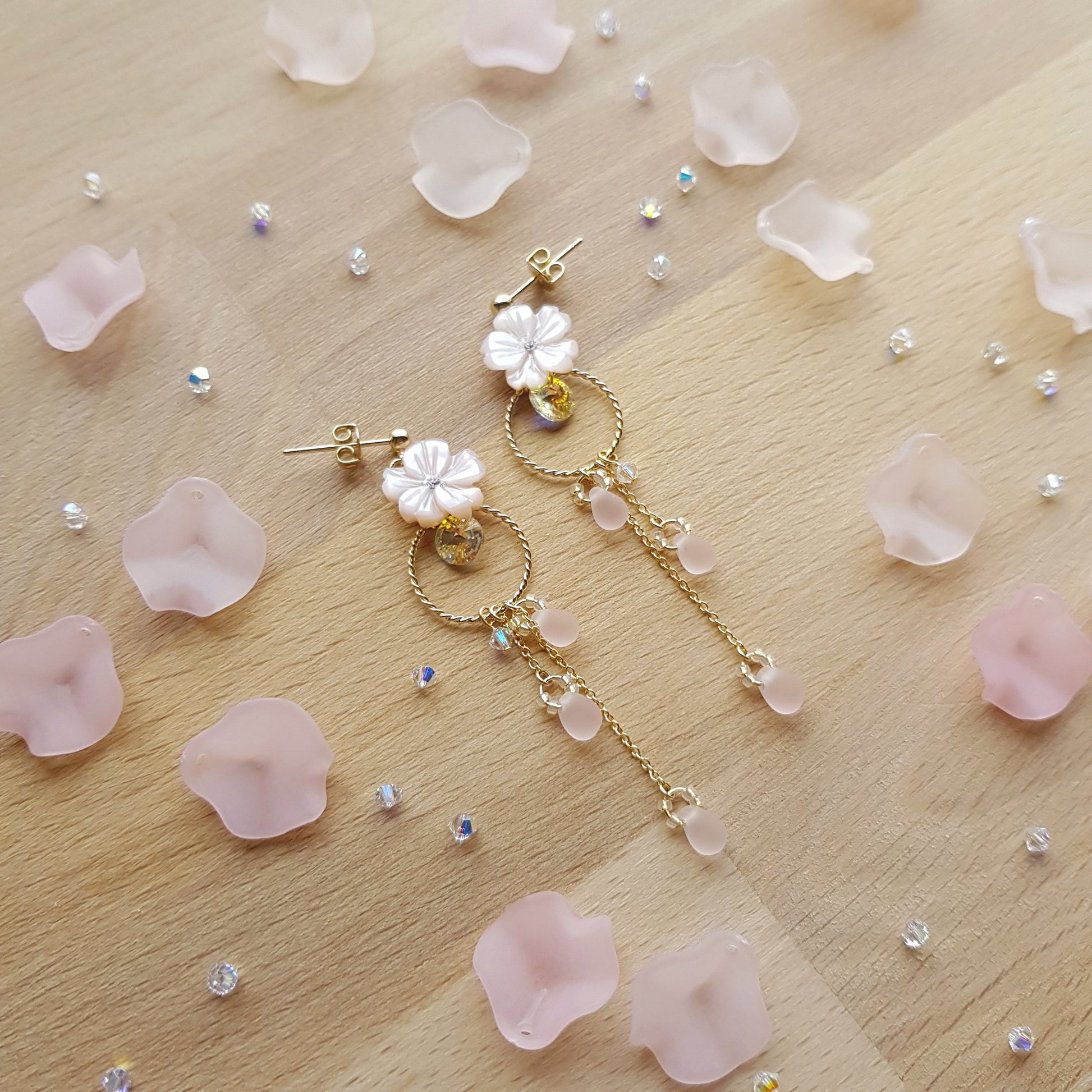 A pair of gold-tone dangle earrings with white flower charms and pink bead accents, arranged on a light wood surface scattered with pale pink petals and small rhinestones.