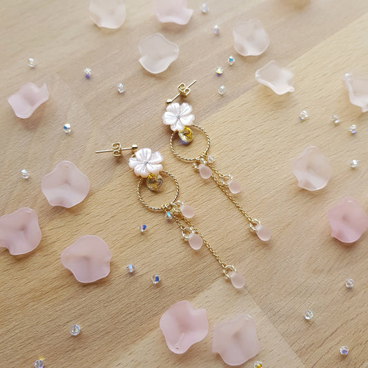 A pair of gold-tone dangle earrings with white flower charms and pink bead accents, arranged on a light wood surface scattered with pale pink petals and small rhinestones.