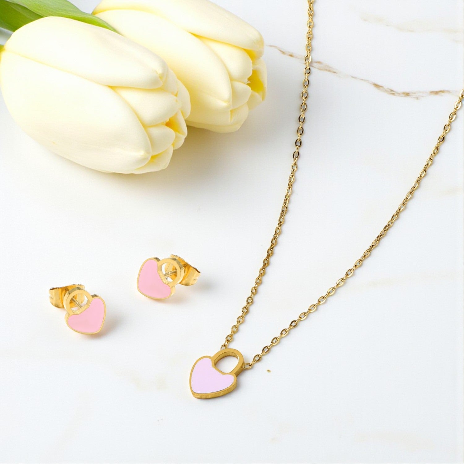 Gold necklace with pink heart pendant and matching earrings on a white surface with tulips.