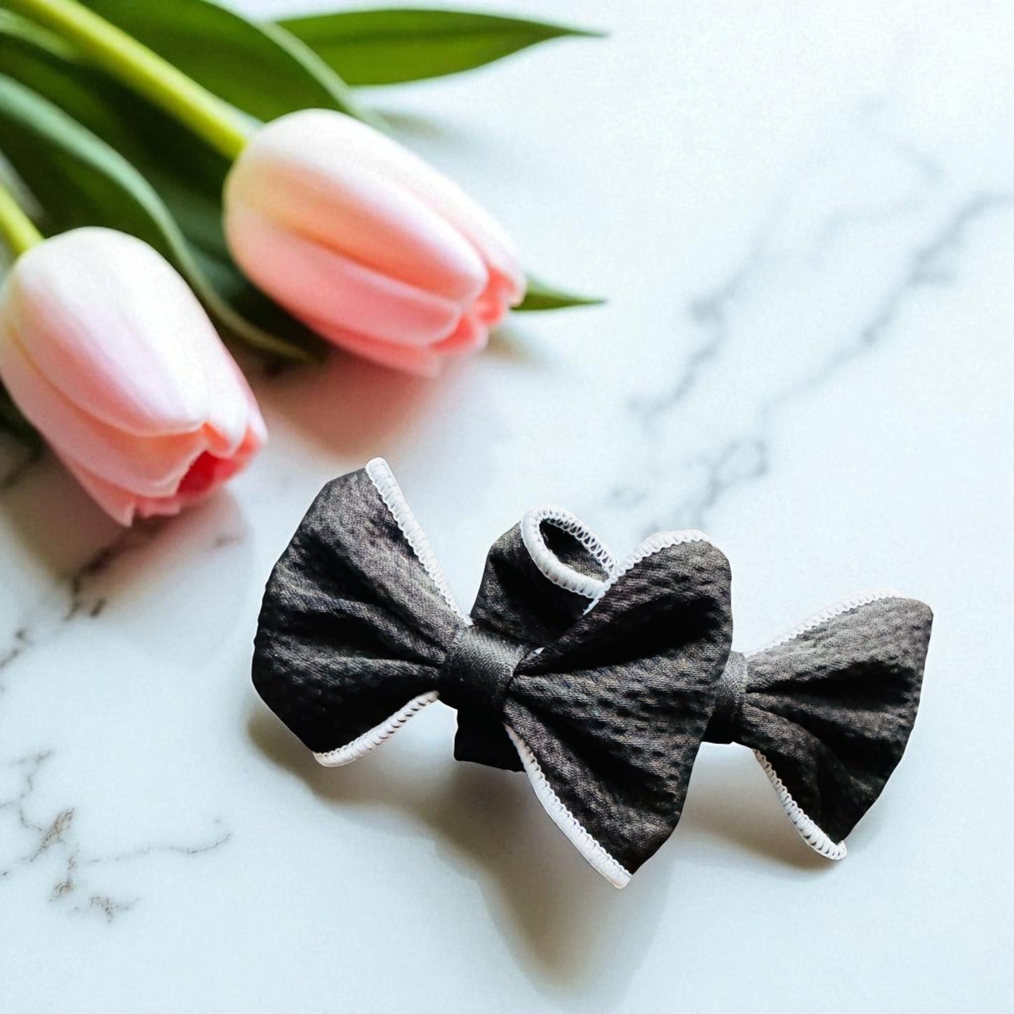 Two gray bow-shaped hair accessories on a marble surface with pink tulips in the background.