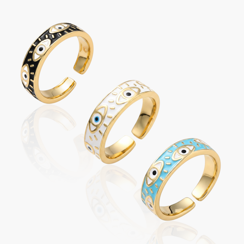 Three gold-tone adjustable ring bands with evil eye enamel details, displayed diagonally on a light gray reflective surface. One ring has black and white eye pattern, one has white and blue eye pattern, and one has light blue and white eye pattern.