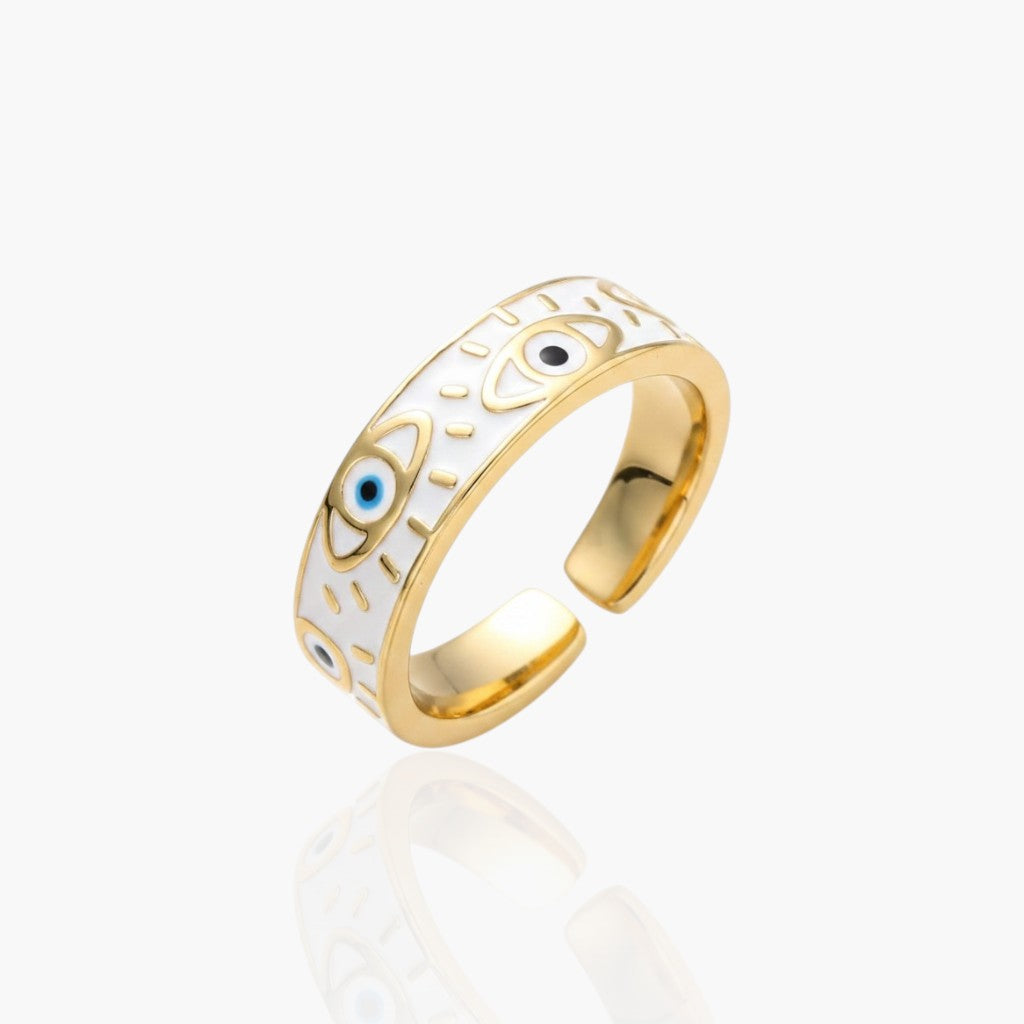 Gold-plated open band ring with white enamel inlay and evil eye motifs, shown on a light gray background with a soft reflection below.