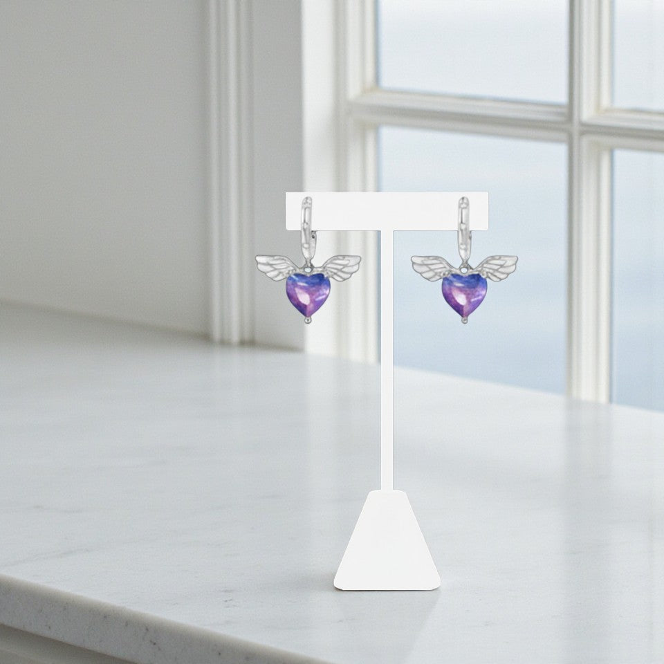 Pair of earrings with purple heart and angel wing design on a white stand against a light background.