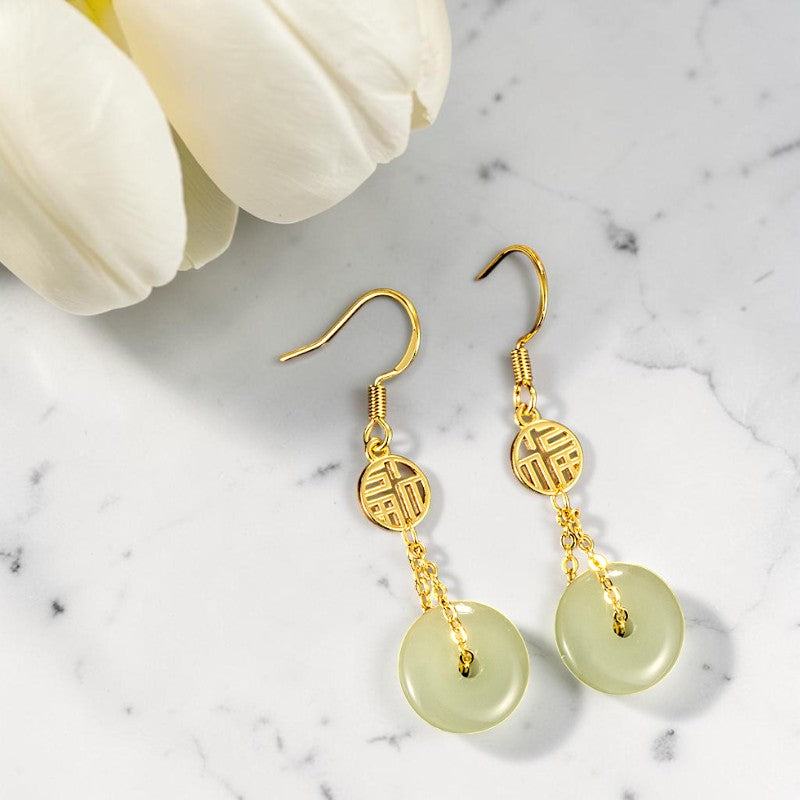 Gold earrings with green circular stones on a marble surface with white flowers.
