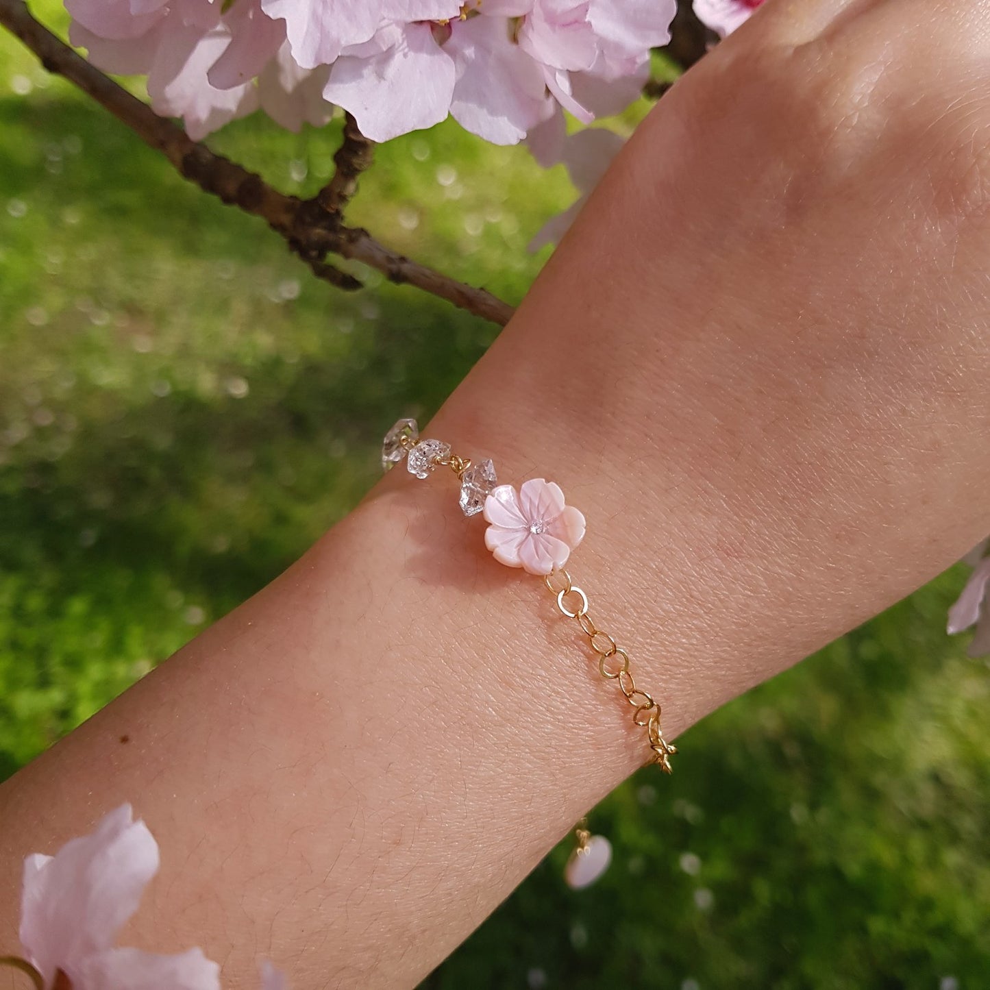 A delicate gold chain bracelet with a pink cherry blossom flower charm and clear crystal accents, shown worn on a person's wrist against a soft-focus background of green grass and pink blossoms.