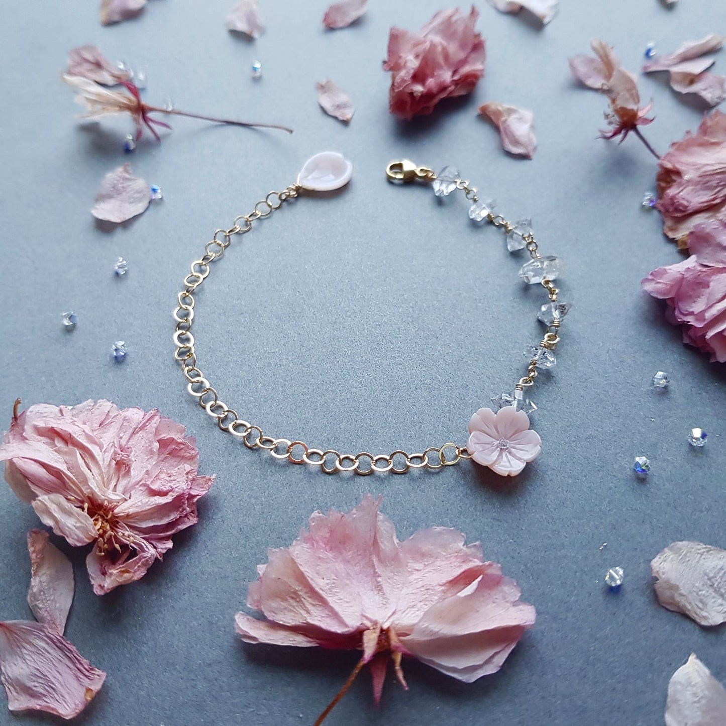 A delicate gold chain bracelet with a light pink cherry blossom flower charm and small clear faceted beads, arranged on a textured blue-gray surface surrounded by pink dried flower petals.