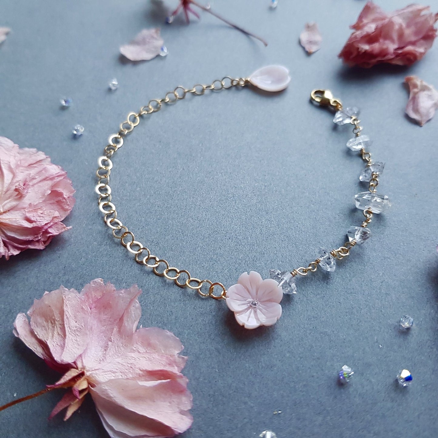 A delicate gold chain bracelet featuring a central light pink cherry blossom flower charm and clear faceted crystal beads, displayed on a textured gray background with scattered pink flower petals.