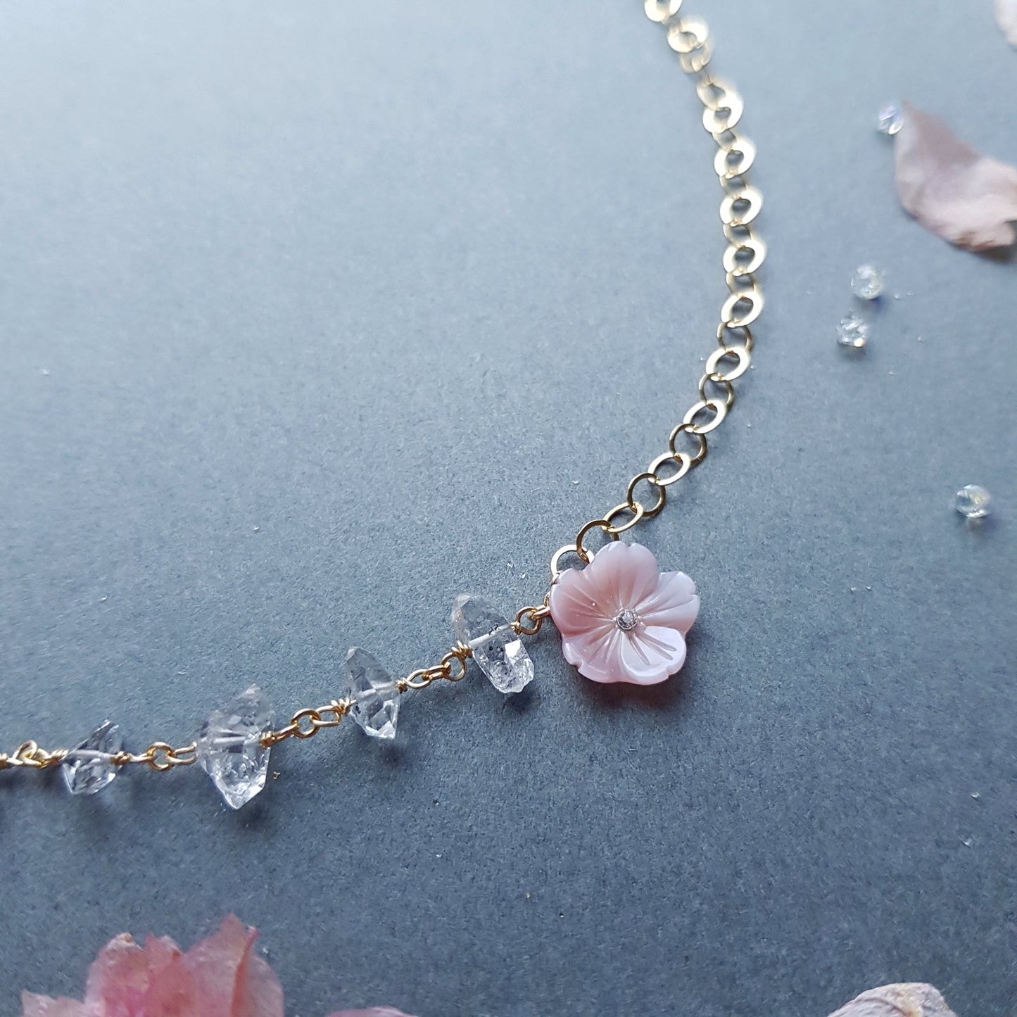 A delicate gold chain necklace featuring a central light pink cherry blossom flower charm and small clear faceted crystal beads, displayed on a textured dark gray surface with scattered pink petals.