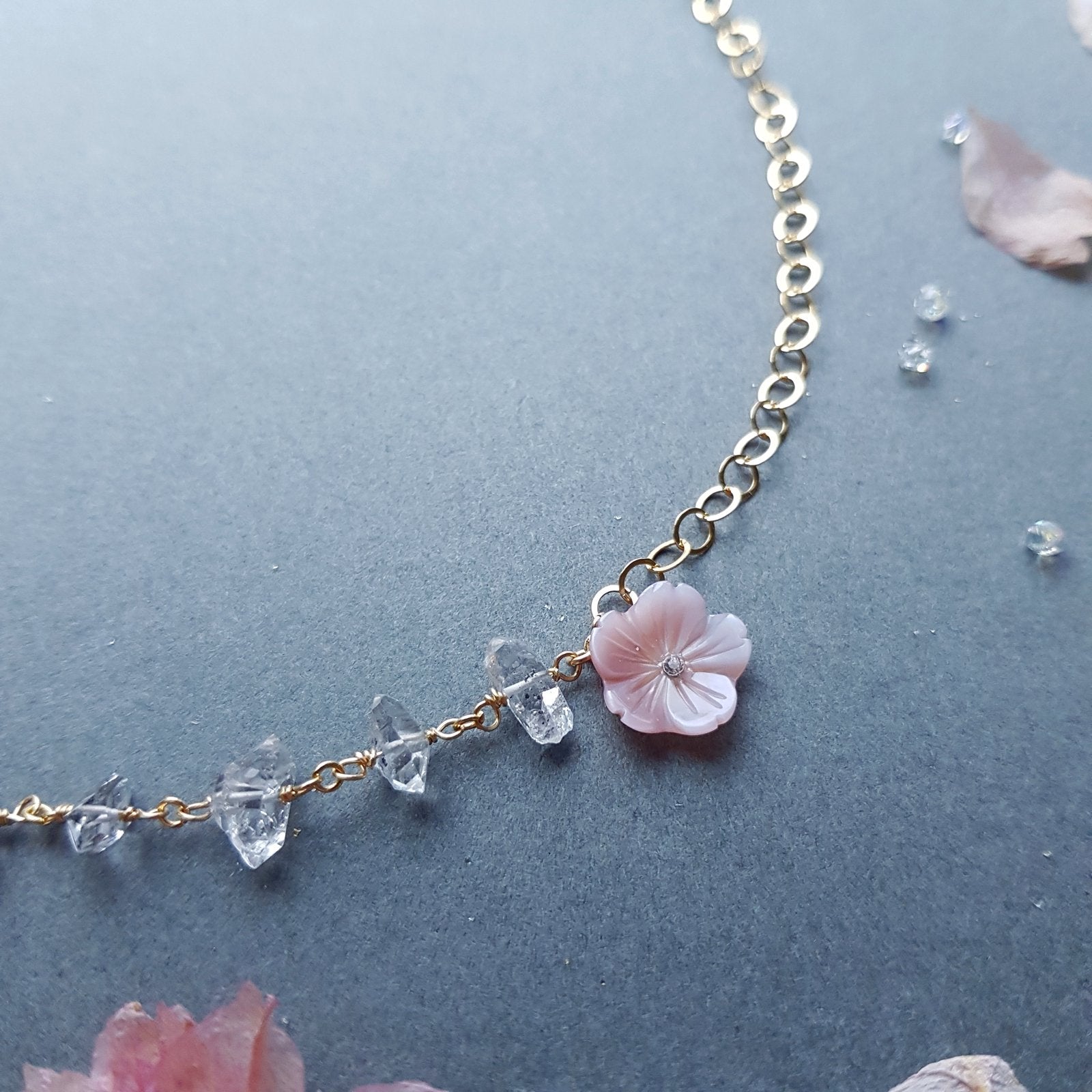A delicate gold chain necklace featuring a central light pink cherry blossom flower charm and small clear faceted crystal beads, displayed on a textured dark gray surface with scattered pink petals.