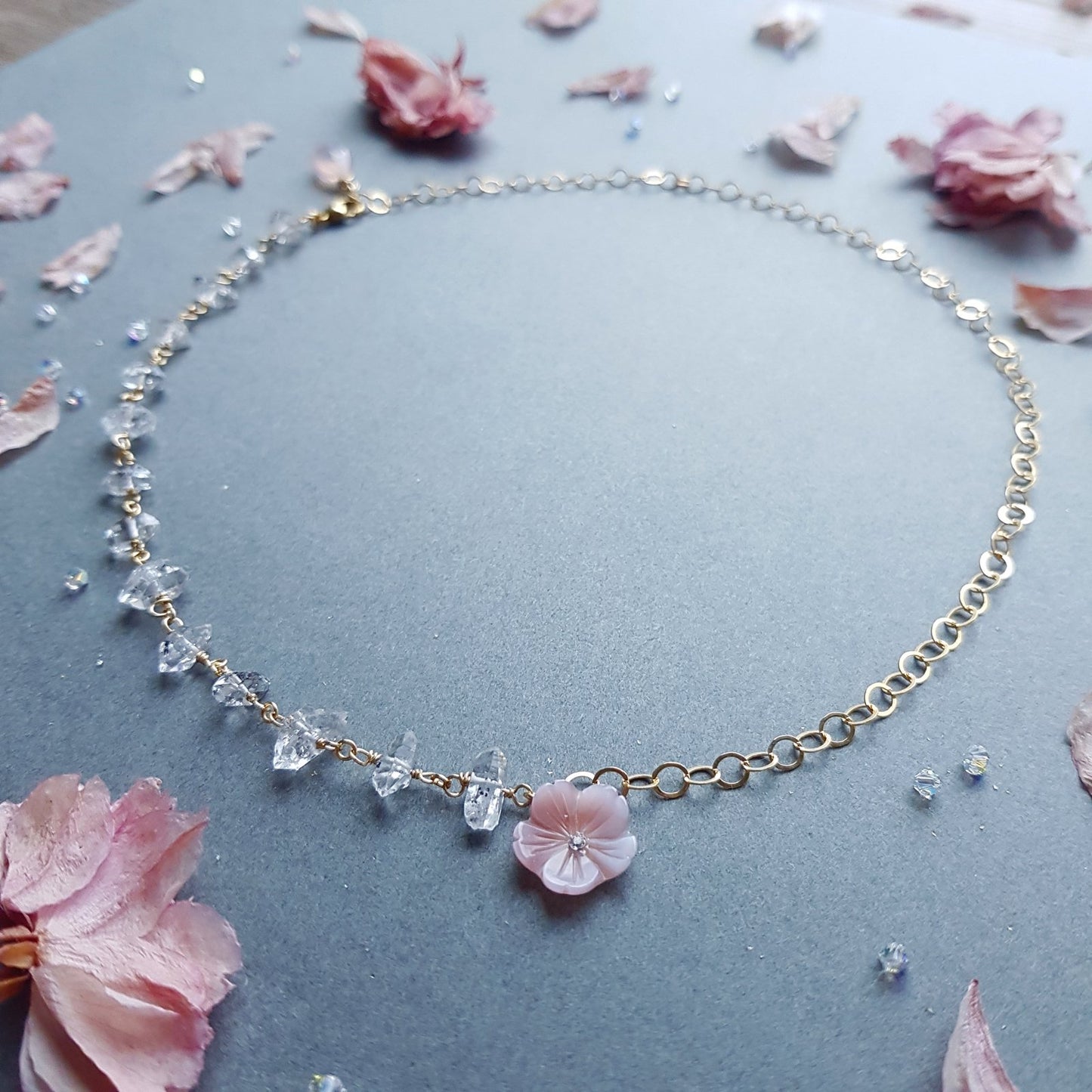 A delicate gold chain necklace featuring clear faceted crystal beads and a small pink flower charm, displayed on a textured gray surface with scattered pink petals.