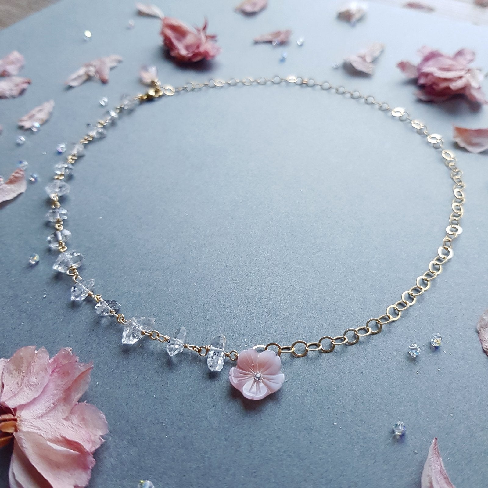 A delicate gold chain necklace featuring clear faceted crystal beads and a small pink flower charm, displayed on a textured gray surface with scattered pink petals.