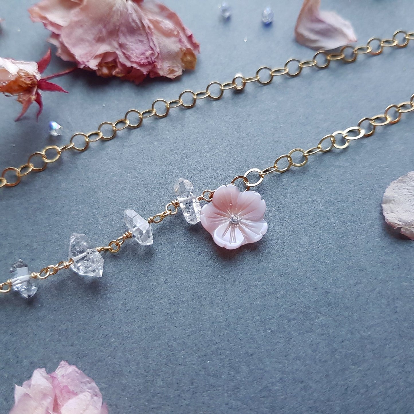 A delicate gold chain necklace featuring a central light pink cherry blossom flower charm and small clear faceted beads, displayed on a textured gray background with scattered pink petals.