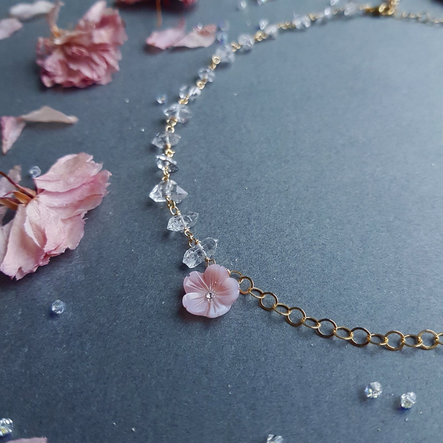 A delicate gold chain necklace featuring a light pink cherry blossom flower charm and clear faceted crystal beads, displayed on a dark gray background with scattered pink petals.