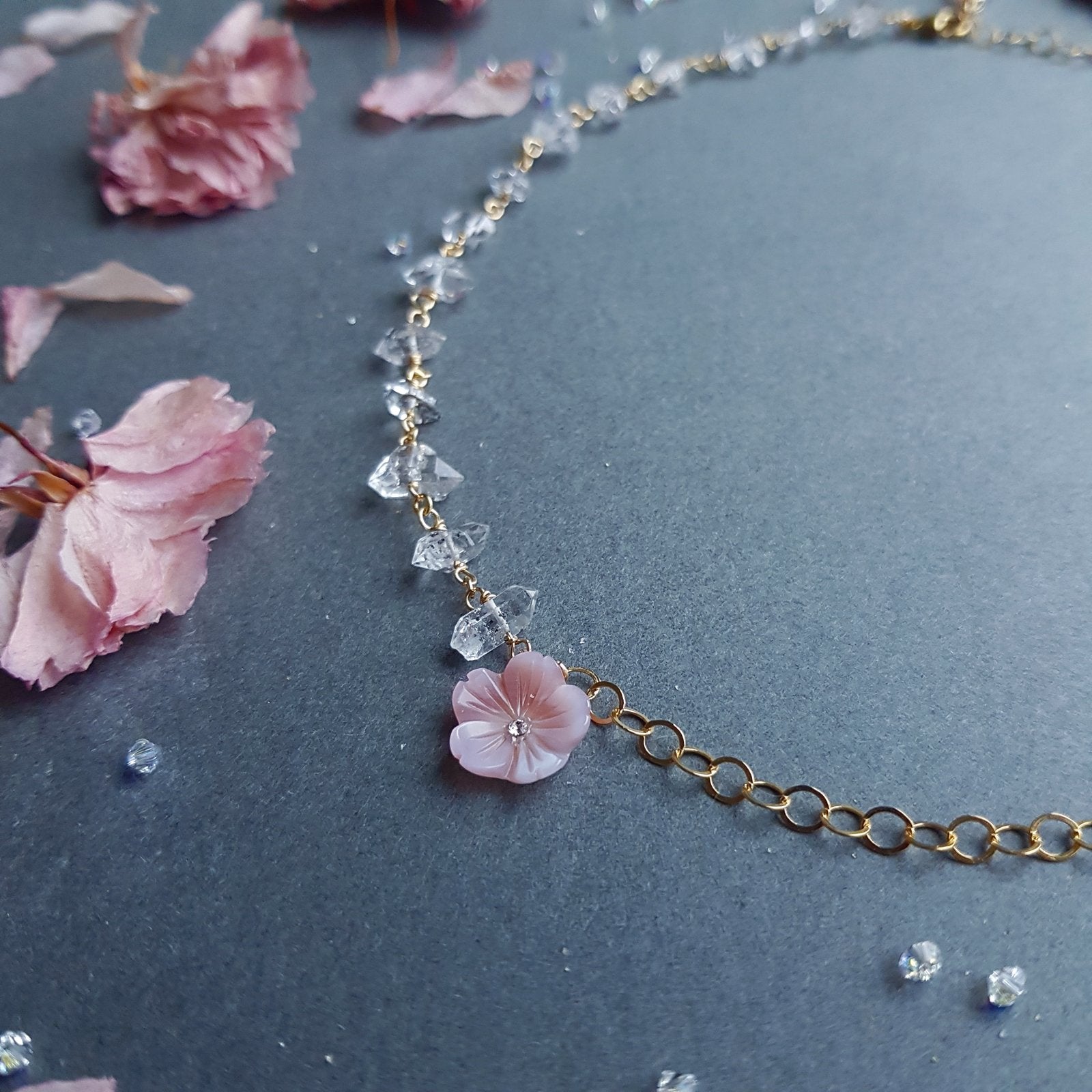 A delicate gold chain necklace featuring a light pink cherry blossom flower charm and clear faceted crystal beads, displayed on a dark gray background with scattered pink petals.