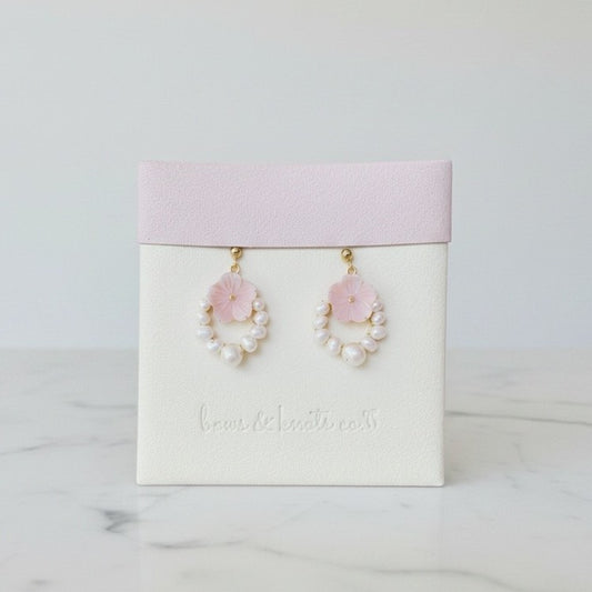 A pair of dangling earrings displayed on a white jewelry card with a pale pink top band. Each earring has a small pink flower above a ring of white faux pearls, with gold-tone hooks.