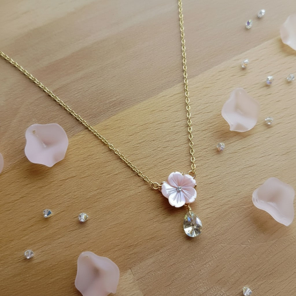 A delicate gold chain necklace featuring a light pink flower charm and a clear teardrop crystal pendant, displayed on a light wood surface with scattered pink petals and small clear stones.