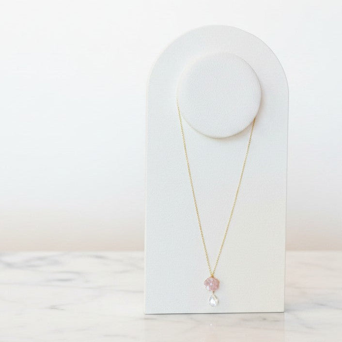A delicate gold necklace with a small pink flower charm and a clear teardrop crystal pendant, displayed on a white arched jewelry stand on a light marble surface.