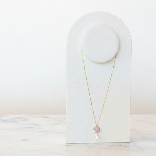 A delicate gold necklace with a small pink flower charm and a clear teardrop crystal pendant, displayed on a white arched jewelry stand on a light marble surface.