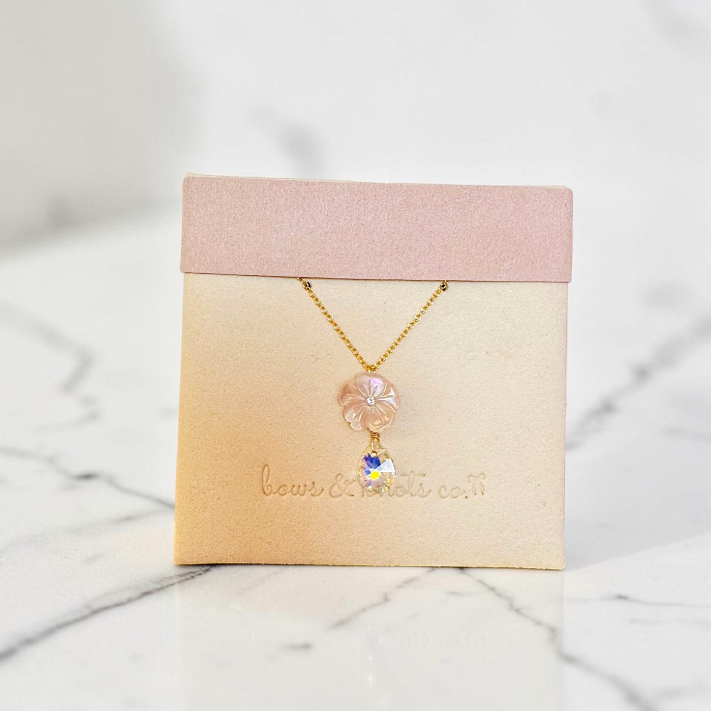 A delicate gold necklace with a pink flower charm and a small white daisy pendant, displayed on a beige jewelry card with a pink top band, set on a white marble surface.