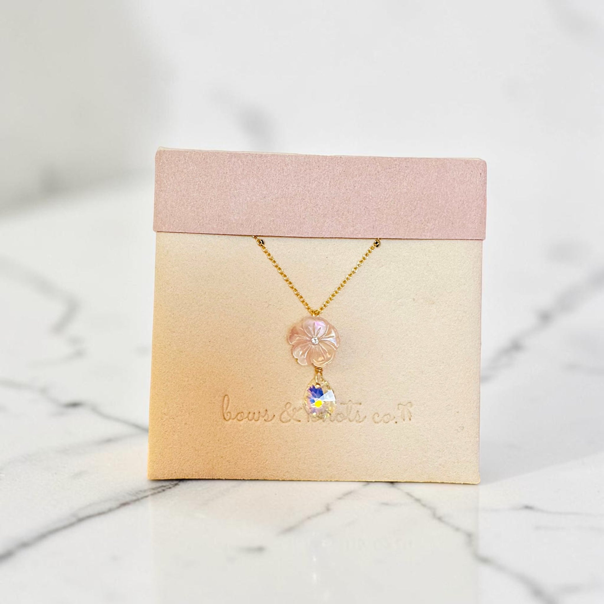 A delicate gold necklace with a pink flower charm and a small white daisy pendant, displayed on a beige jewelry card with a pink top band, set on a white marble surface.