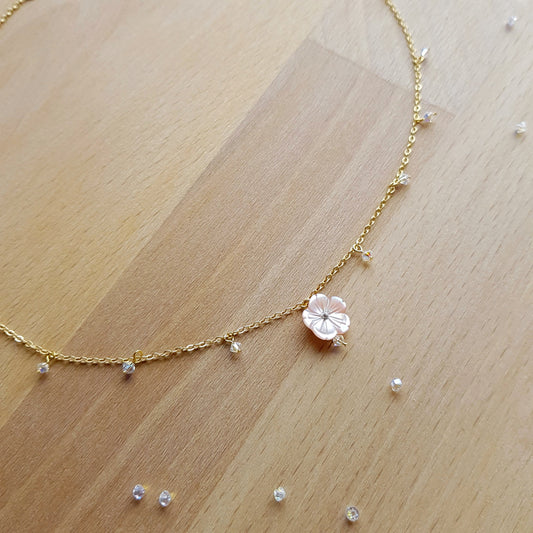 A delicate gold chain necklace with a small pink flower charm and tiny clear stones, displayed on a light wood surface.