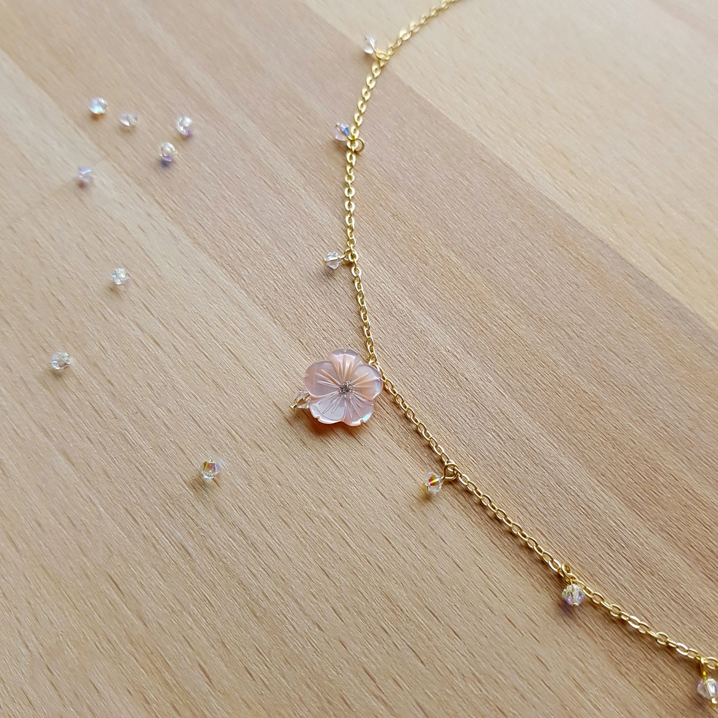 A delicate gold chain necklace with a central pale pink flower charm and small clear crystal beads, laid out on a light wood surface.
