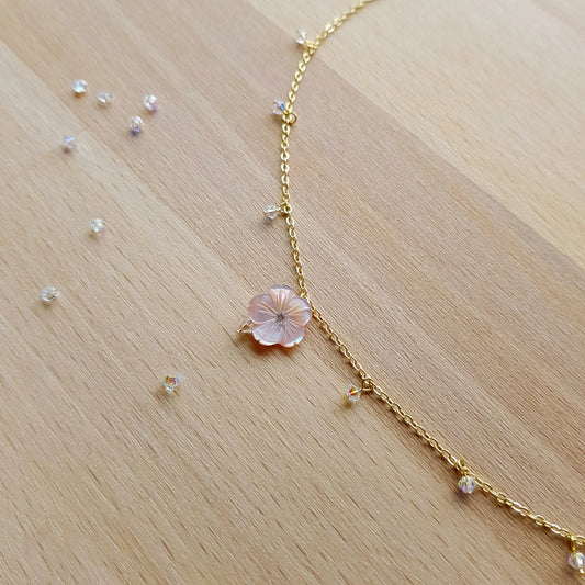 A delicate gold chain necklace with a central pale pink flower charm and small clear crystal beads, laid out on a light wood surface.