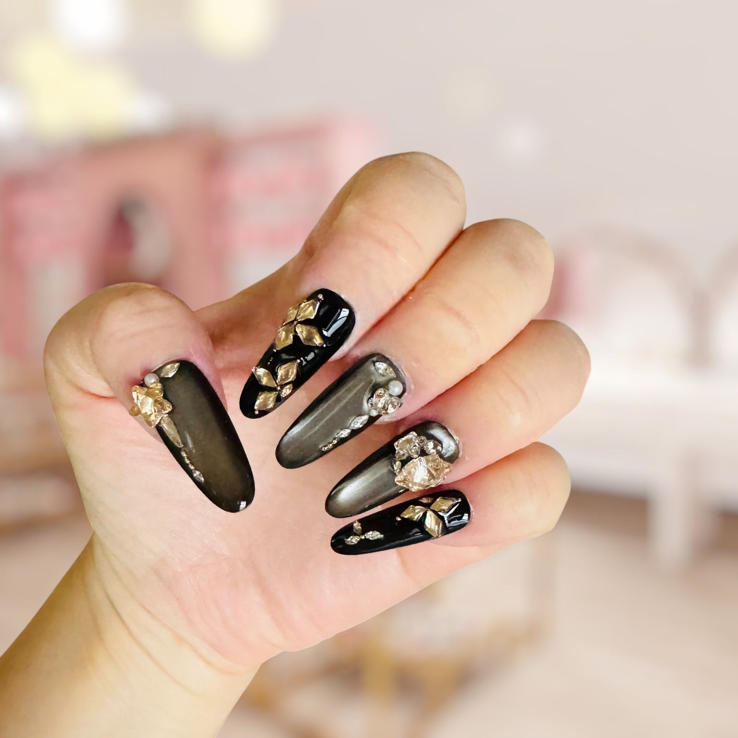 Hand with black nails featuring gold and pearl decorations against a blurred background