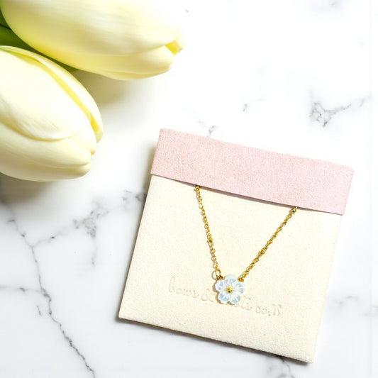 Gold necklace with a flower pendant on a pink and beige card, surrounded by yellow tulips on a marble surface.